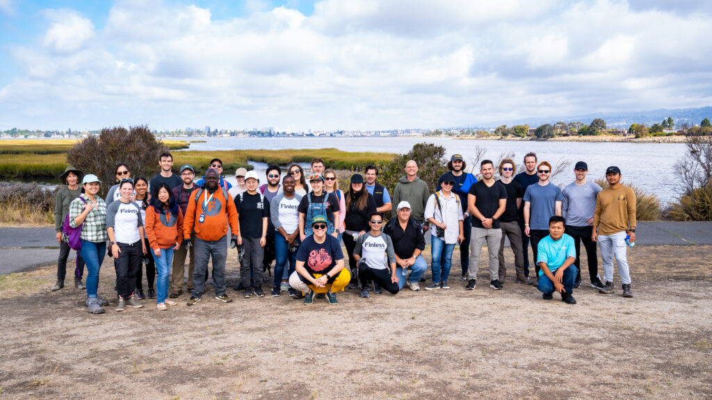 Image description: about 30 people standing and smiling together on a dirt surface in front of a grassy wetland. It is a sunny day with blue skies and some white clouds in the background. 